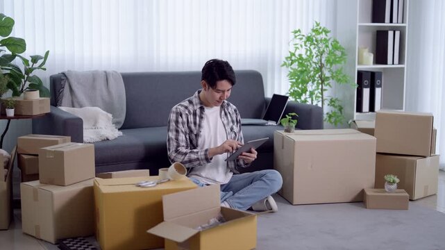 Young Asian man managing online business orders with tablet surrounded by shipping boxes in modern apartment
