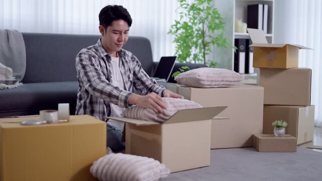 Young Asian man packing cushions into cardboard boxes during home relocation in bright modern apartment