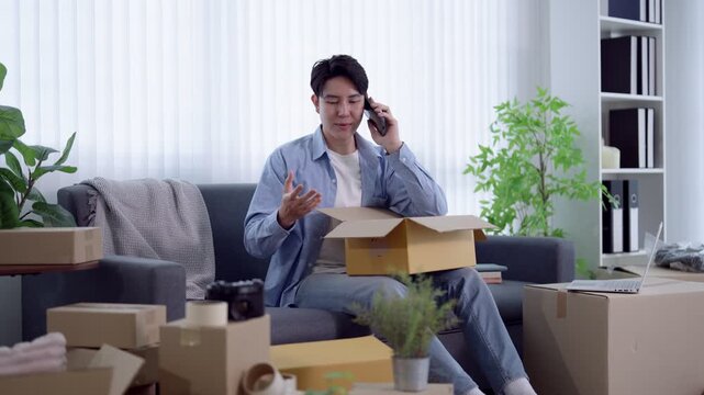 Young Asian man talking on phone while unpacking cardboard boxes in modern apartment with bright natural light