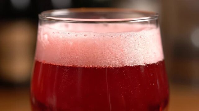 Close-up macro shot of vibrant red or pink sparkling alcoholic or non-alcoholic beverage with thick frothy foam head and rising carbonation bubbles in a glass