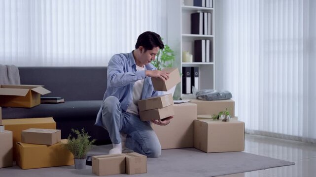 Young Asian man organizing cardboard boxes while kneeling on floor during home relocation with copy space