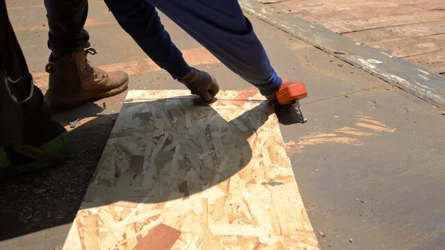 Construction worker installing roofing underlayment on a roof deck using OSB sheathing and a utility knife.