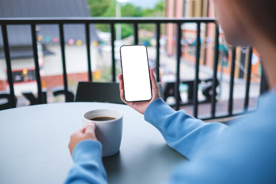 Mockup image of a woman holding and using mobile phone with blank desktop screen while drinking coffee in cafe