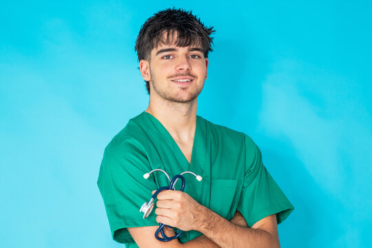 Young doctor or nurse in green surgical scrubs holding a stethoscope, arms crossed, smiling, isolated on a blue background