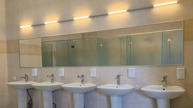 Row of ceramic pedestal sinks with stainless steel faucets and automatic soap dispensers in a modern public restroom. Emphasizing hygiene and accessibility