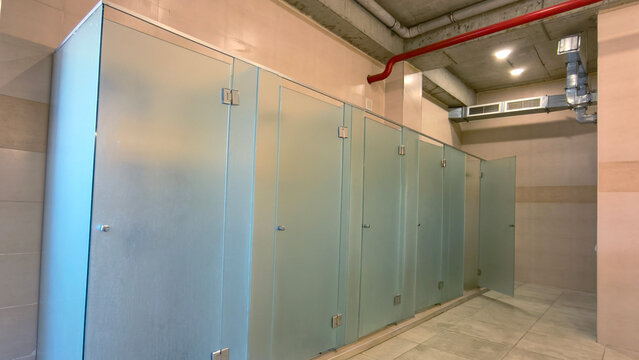 Frosted glass stalls line a public restroom, providing privacy within a clean and modern facility featuring concrete ceilings, exposed pipes, and ventilation systems