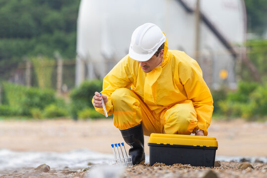 Environmental scientist in hazmat suit and hard hat testing water sample at beach, Researcher in protective suit collecting water quality sample near industrial plant