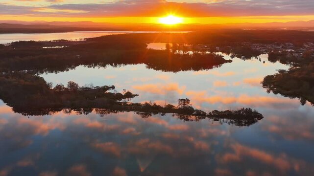 Aerial video gliding over a calm bay as the colours of the sunset sky are reflected across the glassy water, creating a peaceful coastal scene with soft light and tranquil evening conditions.