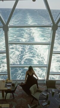 Woman with bare back standing with arms on railing at building window overlooking ship wake; quiet contemplation.