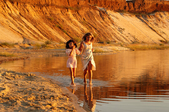 Mother And Daughter Wading Along Sandy Shore At Sunset