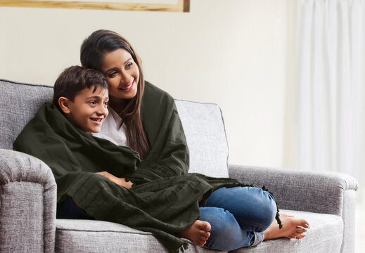 Smiling mother and son wrapped in blanket watching TV on living room sofa