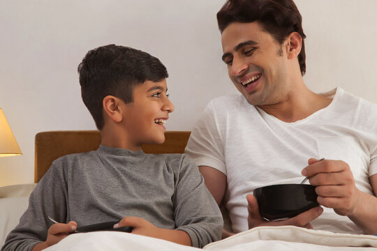 Smiling father and son in bed eating breakfast
