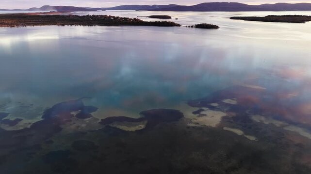 Aerial video gliding over a calm bay as the colours of the sunset sky are reflected across the glassy water, creating a peaceful coastal scene with soft light and tranquil evening conditions.