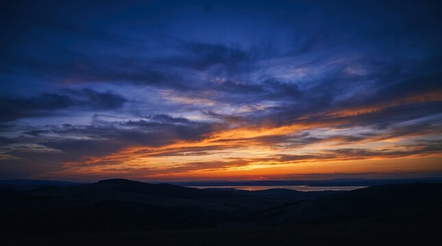 Serene sunset over a calm lake with dark rolling hills and vibrant orange and blue sky