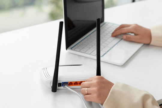 Person connecting an ethernet cable to a white wireless router on a desk with a laptop in the background, a laptop with a person typing on the keyboard is visible in the background, indicating a home