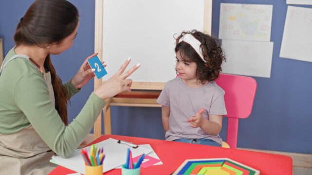 Teacher woman offers numbered flashcards and clipboard while girl child points index finger to a card by an easel in a classroom building; concentration learning.