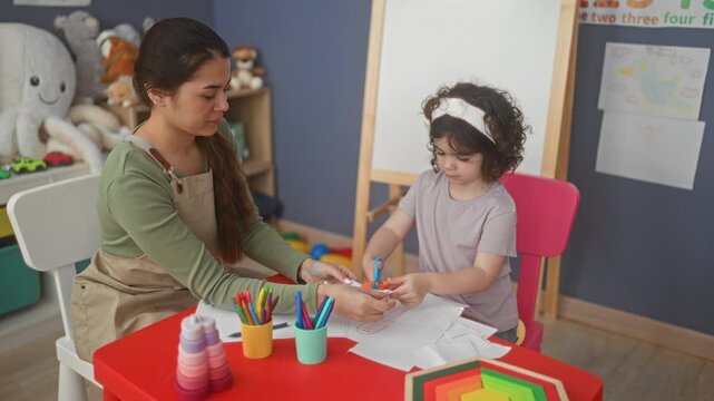 Woman teacher smiles and gives a high five with her hand while guiding a young girl cutting paper with safety scissors at a studio table; learning encouragement.