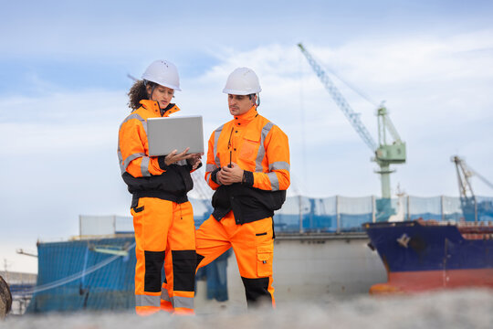 Logistic workers with laptop and radio standing at port shipping yard, Engineers in safety gear discussing cargo management by ship dock, Industrial worker team inspecting logistic operations outdoors