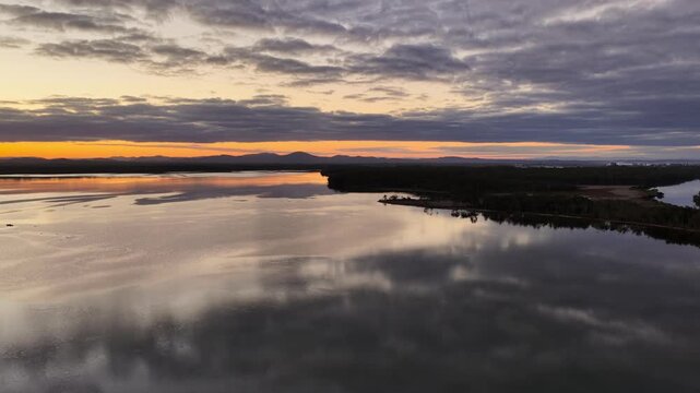 Aerial video gliding over a calm bay as the colours of the sunset sky are reflected across the glassy water, creating a peaceful coastal scene with soft light and tranquil evening conditions.