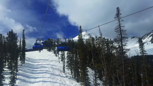 Tilt down handheld winter shot from a ski lift overlooking a Montana mountain landscape with Lone Peak in the distance, snowy trails below, pine forests, and lift chairs passing on a sunny March day.