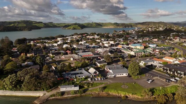 Coastal town spreads across green hillside beside calm estuary at Raglan, New Zealand, helicopter pad and parking lot sit near shops and homes beneath scattered clouds. Aerial drone view.