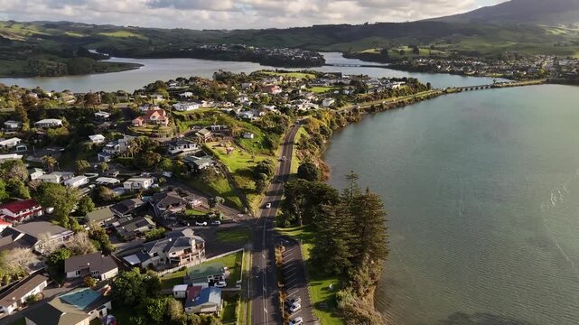Winding coastal road passes waterfront homes and parked cars near estuary shoreline at Raglan, New Zealand, distant bridge and green hills rise beyond calm blue water. Aerial drone wide shot