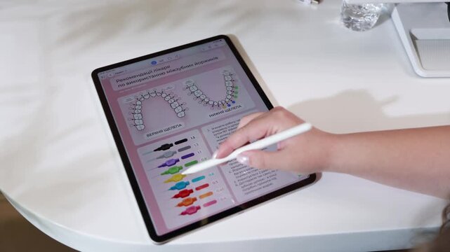 Hand with stylus interacting with tablet showing orthodontic diagram and colorful connector chart on white table in bright clinical workspace, with tablet, stylus, hand, forearm detail