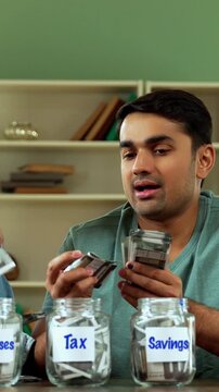 Indian Asian young couple in twenties sitting at home desk, placing Indian currency notes in three glass jars labeled Gold, Mutual Funds, Fixed Deposit, symbolizing savings, investment, financial plan