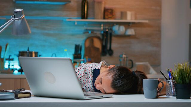Exhausted woman dozes at the kitchen desk beside an open laptop after working overtime on a demanding project late at night.