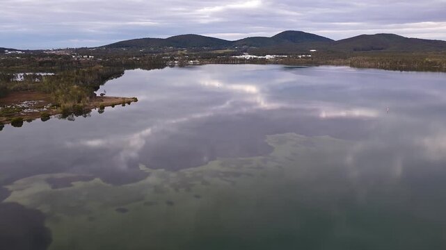 Aerial video gliding over a calm bay as the colours of the sunset sky are reflected across the glassy water, creating a peaceful coastal scene with soft light and tranquil evening conditions.