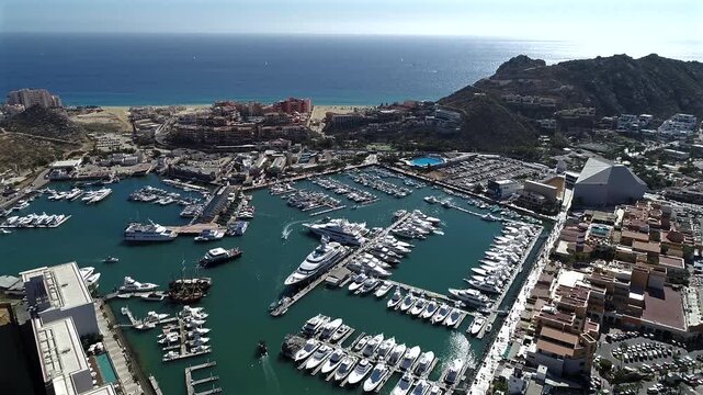 Drone looks down at roughly a 45 degree angle over the Cabo San Lucas marina, moving across a full harbor of yachts and boats, past hotels, and over a luxury beachfront resort.