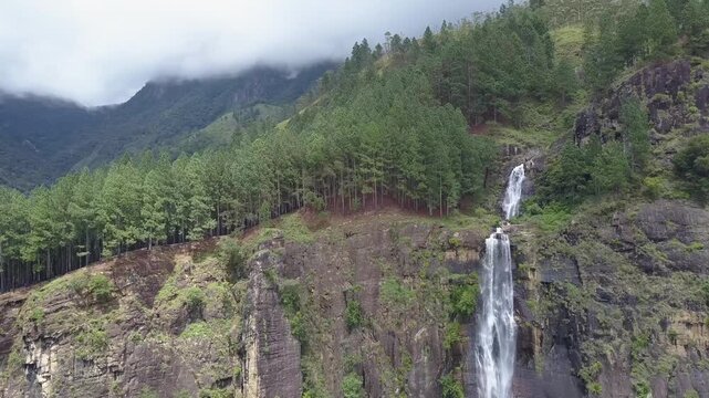 Scenic high aerial view of a dramatic waterfall plunging down a towering rocky cliff framed by pine tree forests and misty green mountains in the highlands of Sri Lanka on a cloudy day.