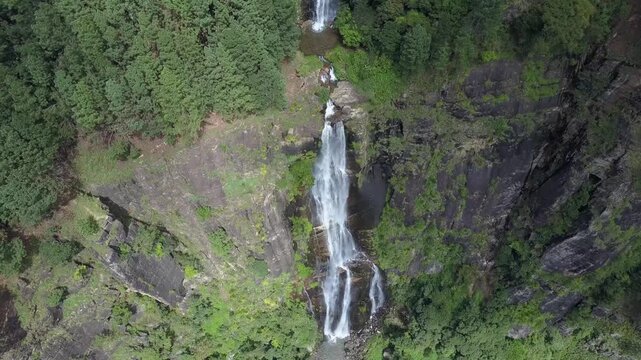 Scenic high aerial view of a dramatic waterfall plunging down a towering rocky cliff framed by pine tree forests and misty green mountains in the highlands of Sri Lanka on a cloudy day.