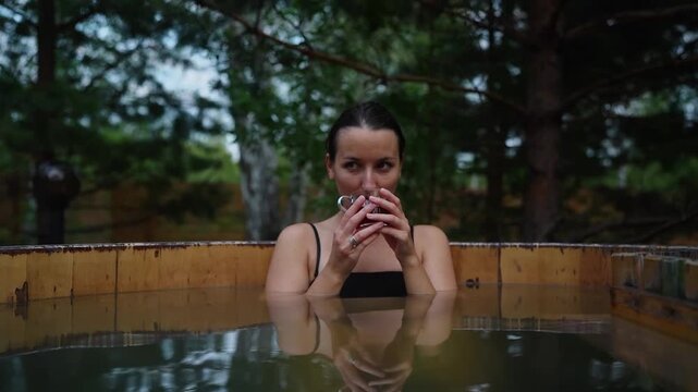 Woman enjoy outside woden japanese steaming hot tub in romantic nature coniferous forest. Idyllic relaxing bathtub in sunset at spa resort in woods nature. Fall winter relax