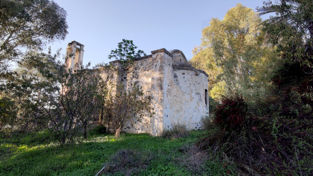Exterior view of the historic Acendu Church (Agios Georgios) in Lefke, showing its stone apse and bell tower surrounded by lush green trees and foliage in warm sunlight.