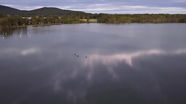 Aerial video gliding over a calm bay as the colours of the sunset sky are reflected across the glassy water, creating a peaceful coastal scene with soft light and tranquil evening conditions.
