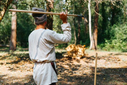A man dressed in simple medieval clothing is poised to throw a spear in a forest clearing. The scene captures the action of a reenactment, highlighting his focused stance and the natural surroundings