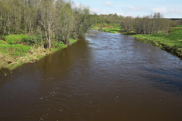 Obraz premium Calm River Flowing Through Spring Countryside with Bare Trees and Green Fields