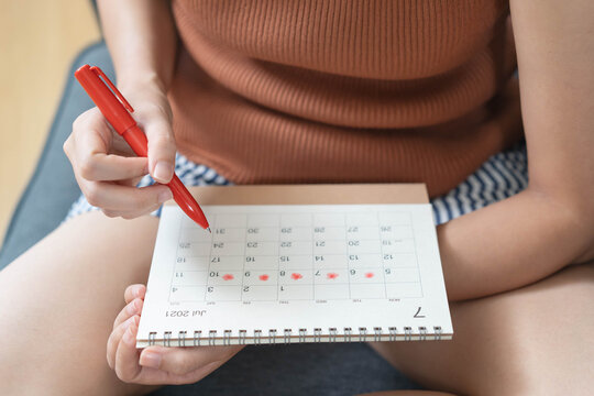 Top view of young woman holding a red pen and tracking her menstrual cycle on calendar, concept of fertility planning, period symptoms and female reproductive health.