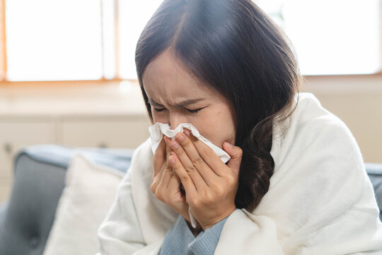 Sick Asian woman blowing her nose with tissue on sofa, wrapped in blanket, suffering from high fever, seasonal flu, influenza virus infection, and cold symptoms at home