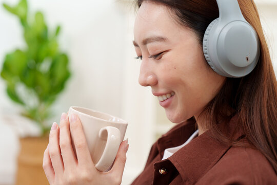 Close up of young, beautiful Japanese woman in brown shirt with headphones holding and looking at coffee or tea mug with both hands, smelling with gentle smile in cozy living room at home