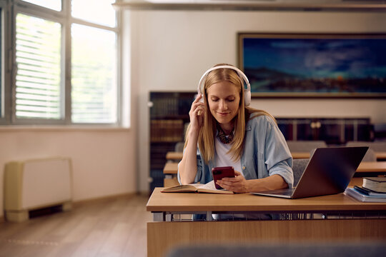 Smiling student using cell phone while wearing headphones in classroom.