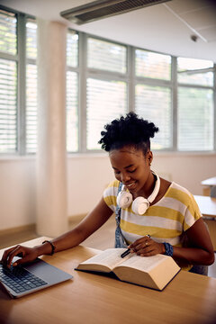Black female student learning from book while using laptop in classroom.