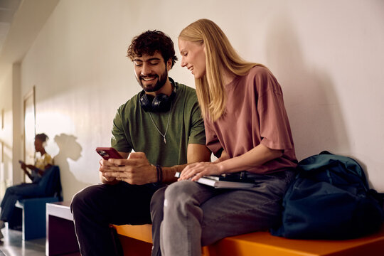 Happy college students using cell phone while sitting in hallway.