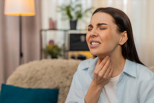Young woman at home sitting on sofa holding throat feeling pain, coughing and looking sick. Caucasian sick girl experiencing flu symptoms, sore throat discomfort, unhealthy, showing illness signs