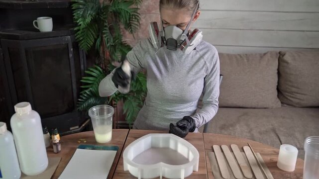 Gloved maker dispensing epoxy resin over heart-shaped silicone mold in cozy home studio workspace with respirator, crafting tools, and soft neutral interior light