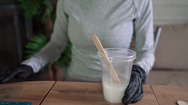 Gloved hands stirring pale epoxy resin in clear mixing cup with wooden craft stick inside bright indoor DIY workspace with plant background and soft light, with glove, stirring, resin, epoxy detail