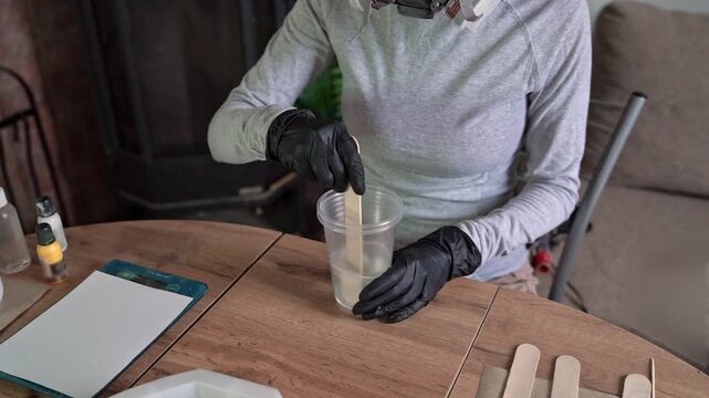 Gloved hands stirring translucent epoxy resin in clear plastic cup over wooden table during crafting project with wooden stick, dark gloves, neutral home workspace