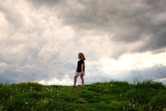 A young girl stands on a hill under dramatic clouds