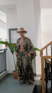 Woman modeling patterned resort wear with bikini top, flowing robe and straw hat in sunlit modern interior.